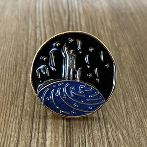 Puede incluir: Un pin de color dorado con un diseño negro y azul. El pin representa dos figuras de pie en un planeta azul con estrellas y estrellas fugaces en el fondo.