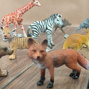 Op de afbeelding: Een verzameling plastic dierenfiguren, waaronder een giraffe, zebra, tijger, vos, beer, nijlpaard, kangoeroe, olifant en een babyolifant. De figuren zijn allemaal in verschillende kleuren en zijn gerangschikt op een houten oppervlak.