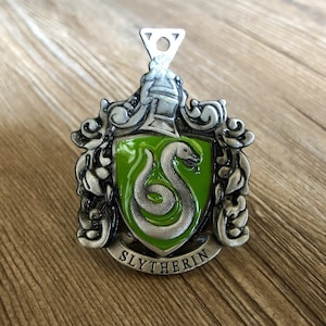 May include: A silver metal charm with a green enamel shield featuring a snake and the words "Slytherin".