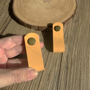 Puede incluir: Dos pequeños tiradores de cajón de cuero naranja claro con remaches color latón. Uno de los tiradores se sostiene en una mano, mientras que el otro está sobre una superficie de madera. El fondo incluye una rodaja de madera y una mesa de madera.