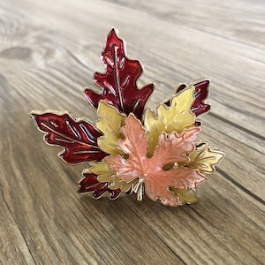 May include: A set of five metal maple leaves in shades of red, orange, and yellow. The leaves are layered on top of each other and have a shiny finish.