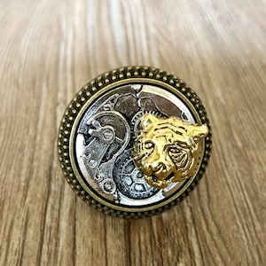 Puede incluir: Un anillo de oro y plata con un diseño de mecanismo de relojería detallado y una cabeza de tigre en la cara.