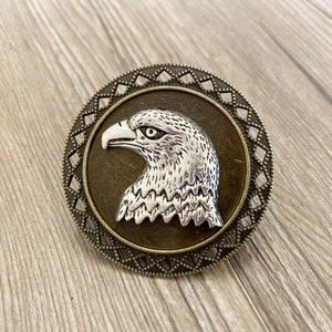 May include: A silver eagle head with a detailed design is featured on a round, bronze-coloured pendant with a decorative border.