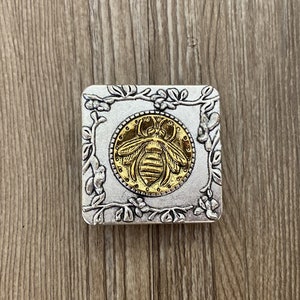 May include: A silver square box with an ornate floral design around the edges. The center of the box features a gold bee with a detailed design.