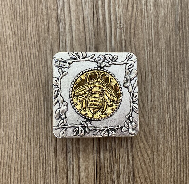 May include: A silver square box with an ornate floral design around the edges. The center of the box features a gold bee with a detailed design.