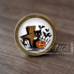 Pode incluir: Um botão de armário redondo de cor dourada com um fundo branco e um gato preto sentado ao lado de uma abóbora de Halloween. O gato está sentado em frente a uma cruz de madeira e uma cerca de metal preto. Há morcegos voando no fundo.