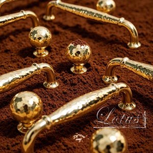 May include: Gold-coloured cabinet hardware, including handles and knobs, with a hammered texture. The hardware is arranged on a brown surface. The word "Lotus" is written in white script.