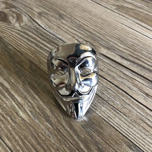 Op de afbeelding: Een zilverkleurig metalen masker met een gestileerd gezicht, dat lijkt op het iconische "V voor Vendetta"-masker.