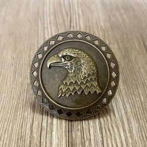 May include: A bronze-coloured metal eagle head ornament with a decorative diamond-shaped border. The eagle's head is facing left and has a detailed design.