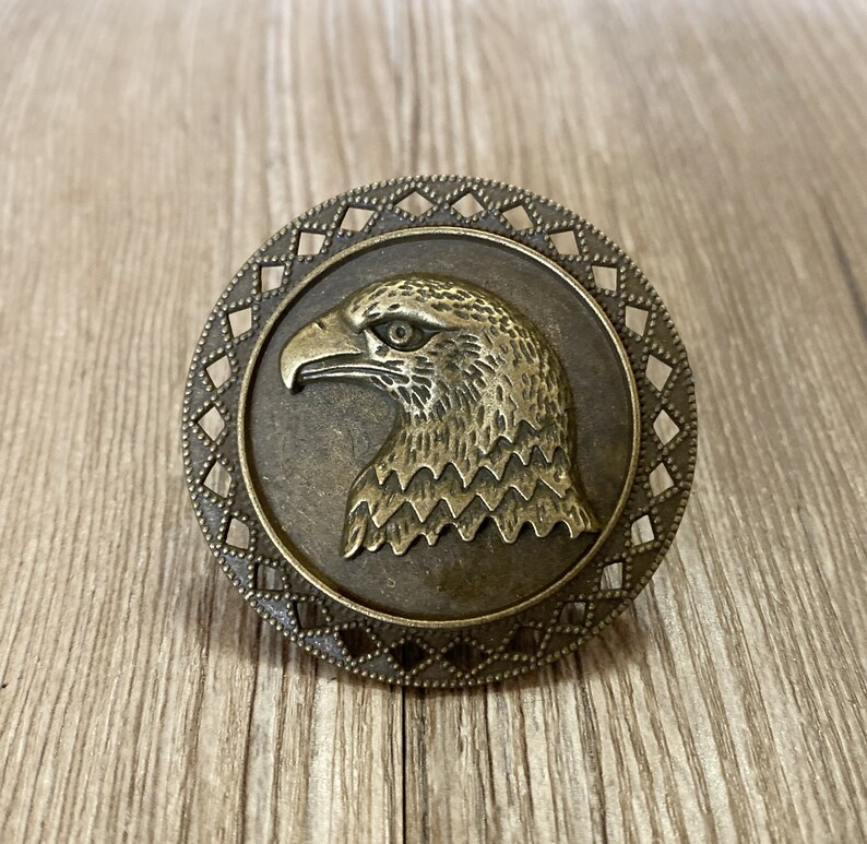 May include: A bronze-coloured metal eagle head ornament with a decorative diamond-shaped border. The eagle's head is facing left and has a detailed design.