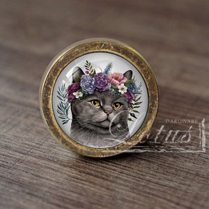 Peut inclure: Bouton de meuble rond avec une finition en laiton. Le bouton présente une illustration détaillée d'un chat gris portant une couronne de fleurs. Les yeux du chat sont jaunes et les fleurs sont violettes, roses et bleues.