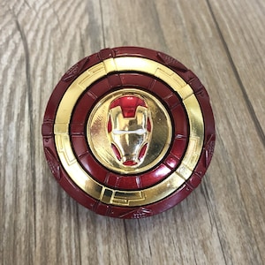 Puede incluir: Un juguete de metal circular rojo y dorado con una imagen en relieve de la máscara de Iron Man en el centro.