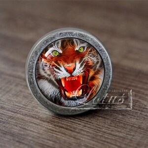 May include: A silver-toned round cabinet knob featuring a tiger face image. The tiger has green eyes and an open mouth with sharp teeth.
