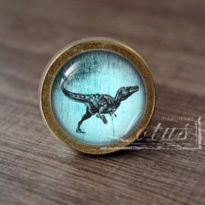 May include: A round, brass-coloured cabinet knob with a blue glass insert featuring a black silhouette of a dinosaur. The knob has a distressed, aged look.