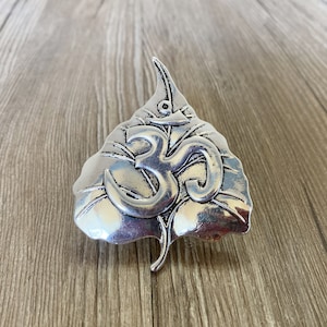 Puede incluir: Un colgante de metal plateado con forma de hoja con el símbolo Om en el centro.