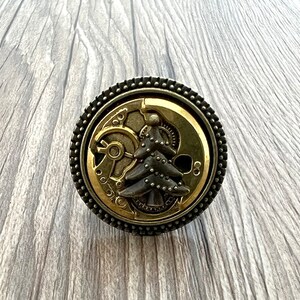 May include: A round, gold-coloured metal knob with a steampunk design. The knob features a stylised tree with gears and other mechanical elements.