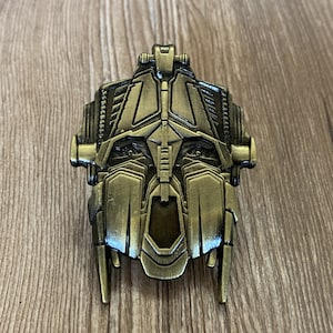 May include: A gold-coloured metal pin featuring a detailed robot face with a menacing expression. The pin has a textured surface and intricate details.