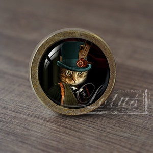 Op de afbeelding: Ronde kastknop met een gedetailleerde afbeelding van een kat met een hoge hoed, een veer en een bloem. De knop heeft een bronzen metalen basis en een glazen afdekking. De kat heeft groene ogen en draagt een zwart pak.