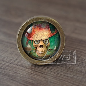 Pode incluir: Puxador de armário redondo em latão antigo com um crânio usando um chapéu de palha. O crânio tem olhos verdes grandes e um sorriso ameaçador. O fundo é uma mistura de verde e castanho, com uma estética vintage.
