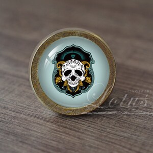 Puede incluir: Un pomo de armario redondo de latón con un inserto de vidrio que presenta una calavera blanca con detalles dorados sobre un fondo azul verdoso y negro.