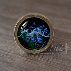 May include: A round, brass-colored cabinet knob with a glass insert featuring a blue and purple seahorse with leafy appendages against a black background.