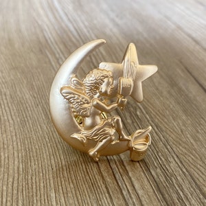 May include: A gold-toned metal pin featuring a cherub holding a paintbrush and sitting on a crescent moon with a star. The cherub is wearing a flowing robe and has wings.