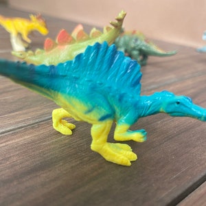 Op de afbeelding: Een verzameling kleurrijk plastic dinosaurus speelgoed. De voorgrond toont een Spinosaurus in blauw en geel. Andere dinosaurussen zijn een gele, groene en blauwe Stegosaurus en andere soorten. Het speelgoed staat op een houten oppervlak.