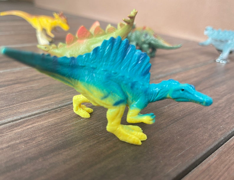 May include: A selection of brightly coloured plastic dinosaur toys. A blue and yellow Spinosaurus is in the foreground. Other dinosaurs include a yellow, green, and blue Stegosaurus and other species. The toys are on a wooden surface.