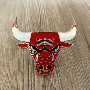 Puede incluir: Un pin de metal de cabeza de toro rojo y blanco con contorno negro. El toro tiene una expresión feroz con nariz y ojos negros.