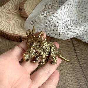 Op de afbeelding: Een gedetailleerd Triceratops beeldje van messing. De dinosaurus heeft een getextureerd oppervlak en staat in een loopstand. Het beeldje is goudkleurig en heeft drie hoorns en een kraag. De achtergrond omvat houten schijfjes en wit kant.