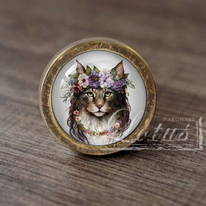 Peut inclure: Un bouton de porte rond en laiton avec un insert en verre représentant un chat portant une couronne de fleurs. Le chat a une fourrure brune et des yeux verts. Les fleurs sont roses, violettes et blanches.