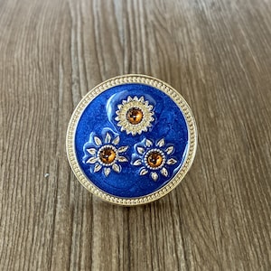 May include: A gold-toned trinket box with a blue enamel lid featuring three stylized sunflowers with orange centers.