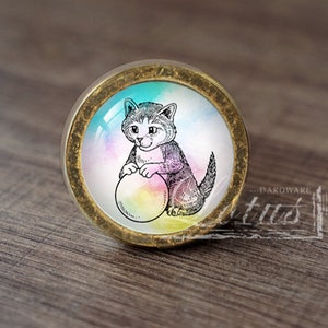 Può includere: Un pomello rotondo per armadio di colore dorato con uno sfondo colorato in stile acquerello. Un'illustrazione in bianco e nero di un gattino che gioca con una palla è centrata sul pomello.