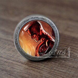 Könnte beinhalten: Ein runder, silberfarbener Metallknopf mit gewölbtem Glaskern. Der Kern zeigt eine rot-orangefarbene Illustration einer Person. Das Wort "Lotus" ist auf einem kleinen, rechteckigen Etikett aufgedruckt.