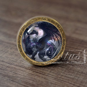 May include: A round, gold-colored knob with a glass insert featuring a black dragon with iridescent wings. The dragon is depicted in a dynamic pose, with its wings spread wide and its head turned to the side. The glass insert is surrounded by a gold-colored metal frame.