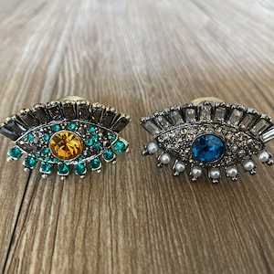 Puede incluir: Dos amuletos decorativos de ojo turco con pedrería y una joya central. Un amuleto tiene un esquema de colores verde y dorado, mientras que el otro tiene un esquema de colores blanco y azul.