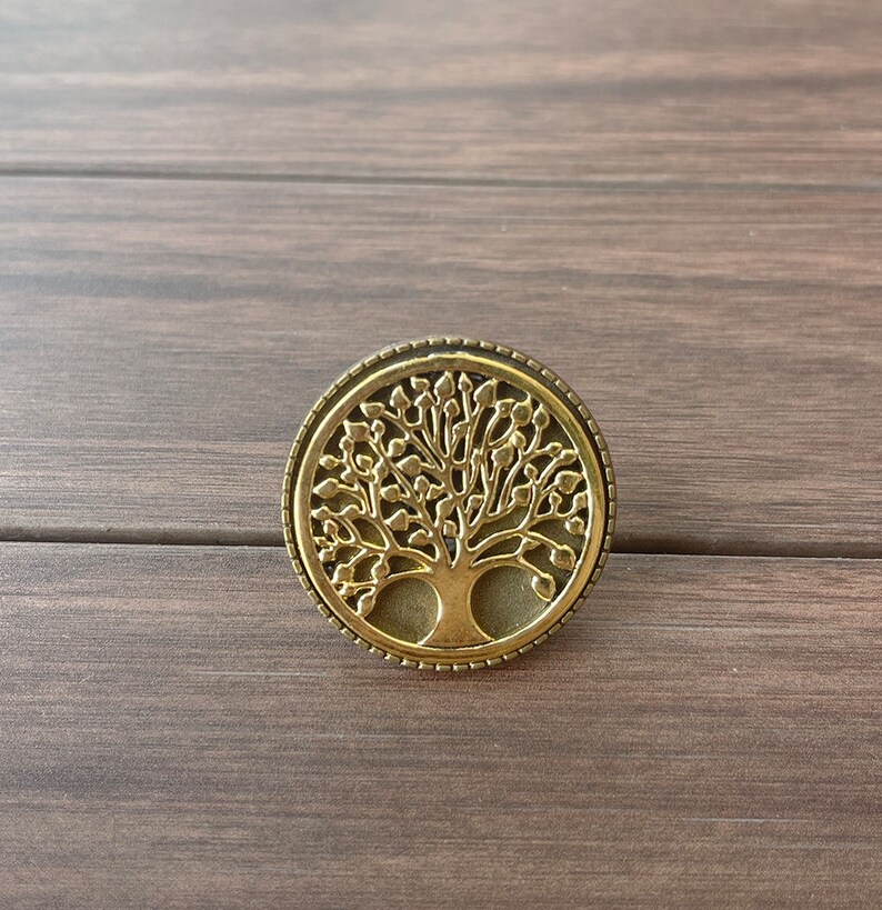 May include: A gold-toned metal pendant featuring a detailed tree of life design. The pendant is circular and has a textured border.