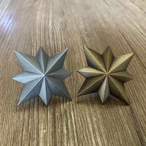 Puede incluir: Dos adornos decorativos en forma de estrella, uno plateado y otro dorado, con una superficie texturizada. Los adornos están sobre una superficie de madera.