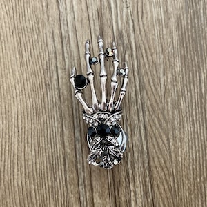 K&ouml;nnte beinhalten: Ein silberner Skeletthand-Anh&auml;nger mit schwarzen und klaren Strasssteinen. Die Hand h&auml;lt eine schwarze Schleife mit Strasssteinen.