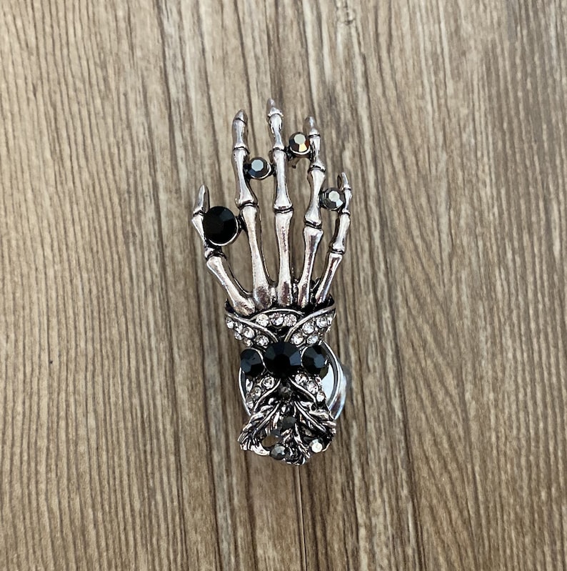 K&ouml;nnte beinhalten: Ein silberner Skeletthand-Anh&auml;nger mit schwarzen und klaren Strasssteinen. Die Hand h&auml;lt eine schwarze Schleife mit Strasssteinen.