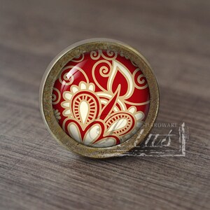 May include: A circular cabinet knob with a red and cream floral pattern, encased in a brass-toned metal frame. The knob has a glossy finish and is set against a wooden surface.