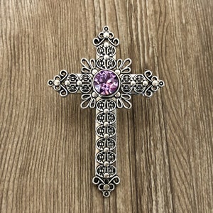 Könnte beinhalten: Ein silbernes Kreuz mit einem verzierten Design und einem violetten Strassstein in der Mitte.