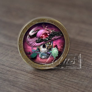 Pode incluir: Um botão de armário redondo de cor latão com um inserto de vidro que apresenta uma caveira usando um chapéu rosa e preto com uma rosa branca na boca. O fundo é um pôr do sol rosa e roxo.