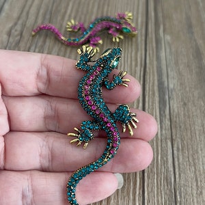 May include: Two jeweled lizard brooches. The front brooch is teal and pink with gold-colored accents. The second brooch is in the background and is pink and teal. The brooches are on a wooden surface.