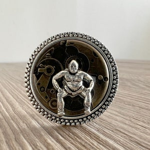 May include: A silver ring with a detailed design of a sumo wrestler in a squatting pose. The wrestler is surrounded by a circular gear-like pattern.