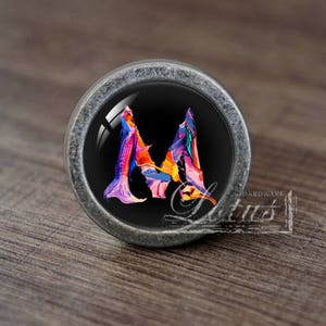 May include: A silver cabinet knob with a black background and a colourful abstract letter M.
