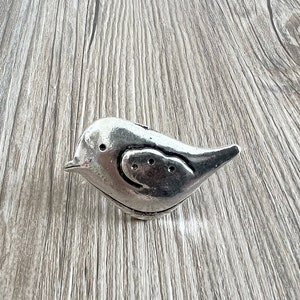 May include: A silver-coloured bird-shaped salt and pepper shaker. The bird has a rounded body, a small beak, and three holes on its side. The shaker is made of metal and has a slightly distressed finish.
