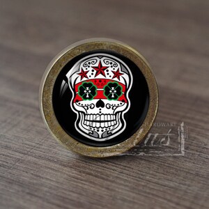 May include: A round, brass-colored cabinet knob with a black background and a white sugar skull design. The skull has red and green details and three red stars above it.