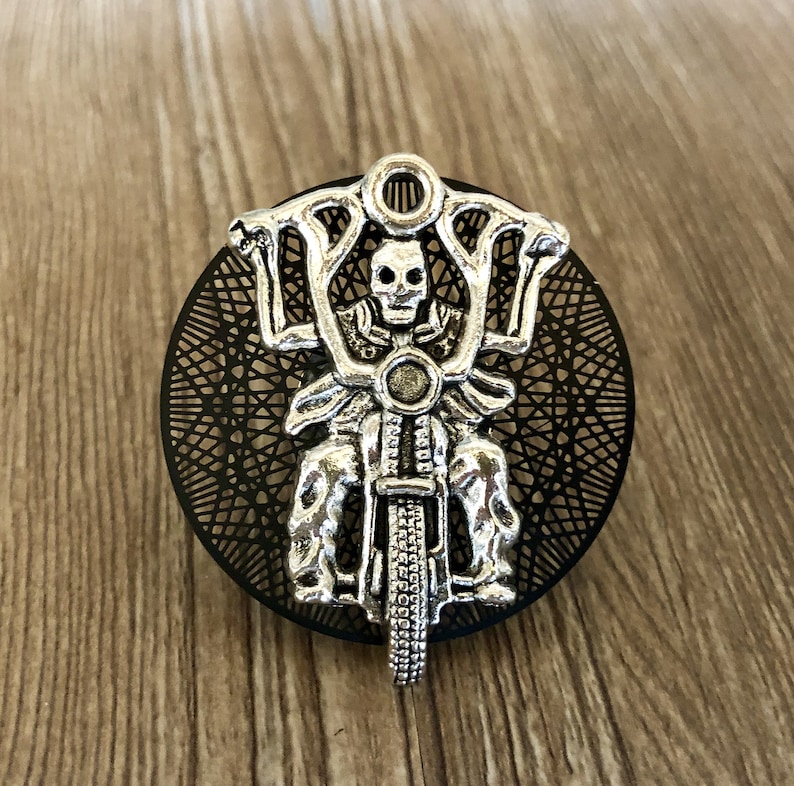 Op de afbeelding: Een zilveren skelet dat een motorfiets bestuurt met een zwart-wit mandala-ontwerp op de achtergrond.