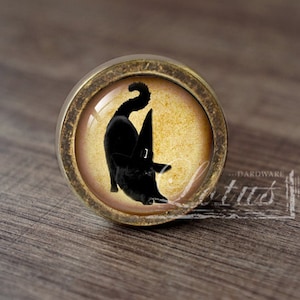 Może przedstawiać: Okrągła gałka do szafki ze starego mosiądzu z czarnym kotem w kapeluszu czarownicy na złotym tle. Ta dekoracyjna gałka to element okuć meblowych.